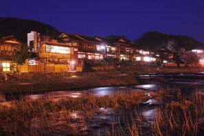 Ett traditionellt japanskt ryokan med flera byggnader i trä, en flod i förgrunden och ett bergigt landskap i bakgrunden.
