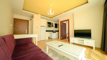 Una sala de estar con sofá, una mesa de centro de vidrio, un televisor sobre un mueble bajo, y una cocina al fondo.