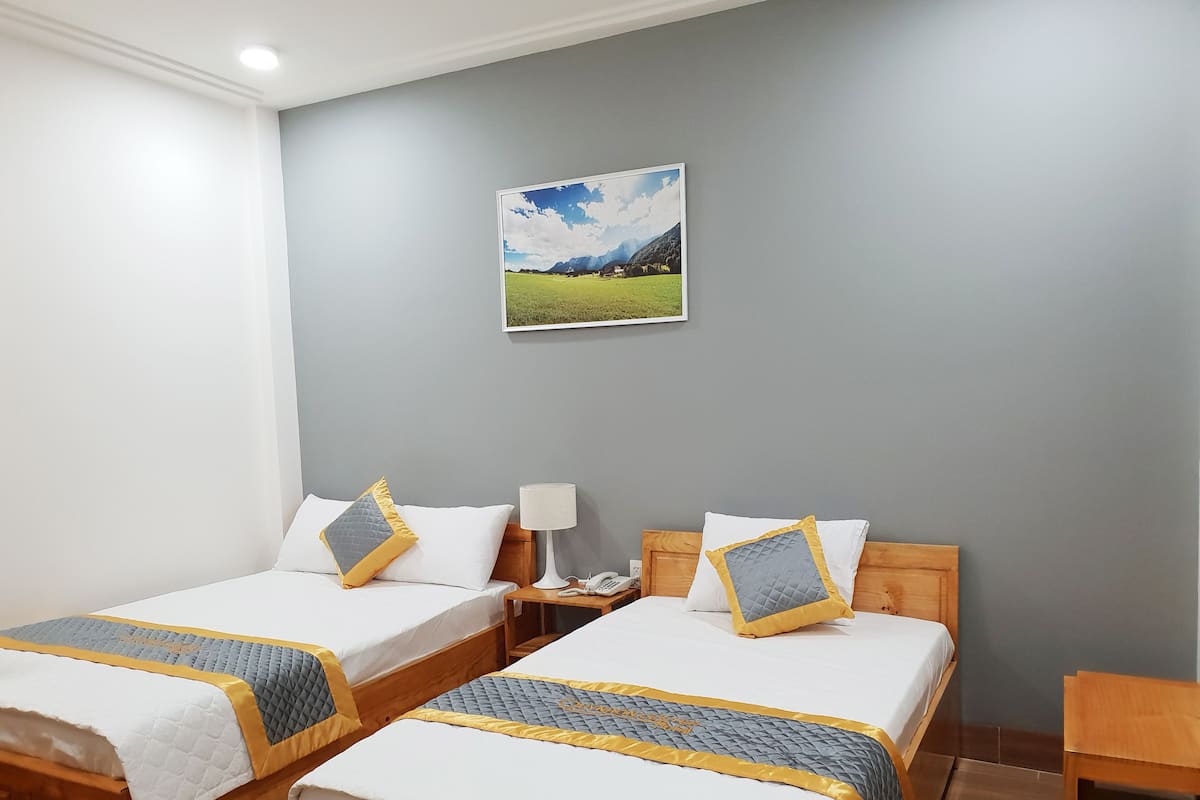 A hotel room with two beds, each with a white and blue quilted bedspread and matching pillows. There is a wooden nightstand with a lamp and a phone, and a framed landscape painting on the wall.