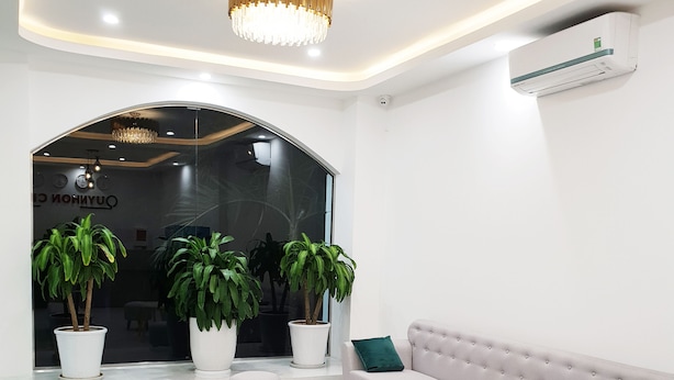 A modern living room with a light grey sofa, two white round tables, and a large chandelier. There are potted plants and a large window in the background. - Khách sạn Quy Nhơn Center (Quy Nhon)