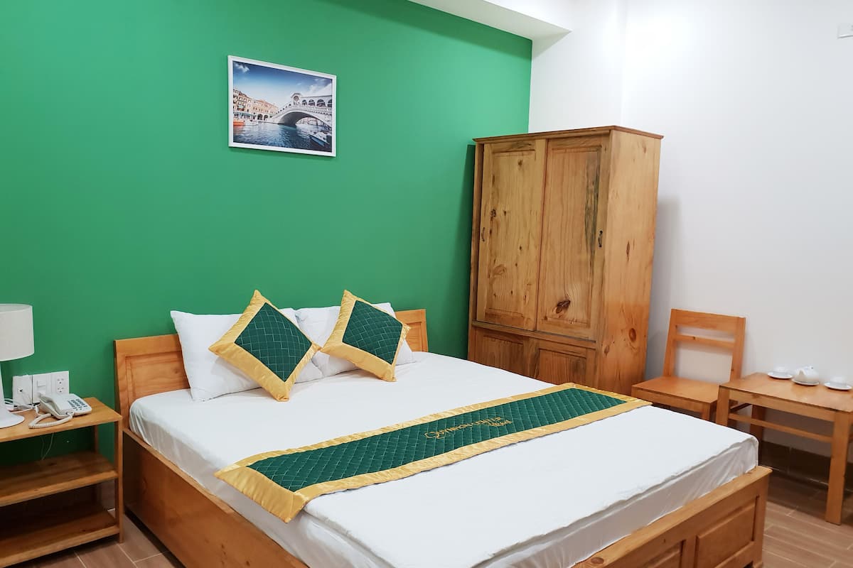 A bedroom with a green wall, a wooden wardrobe, a bed with white bedding and green and gold pillows, a bedside table with a lamp and phone, and a picture of a bridge on the wall.