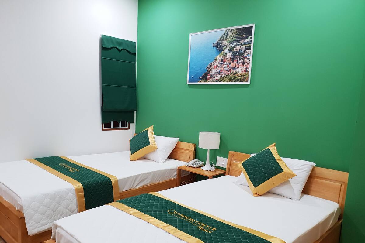 A hotel room with two beds, each with green and gold bedspreads, a nightstand with a lamp, and a framed picture of a coastal village on the wall.