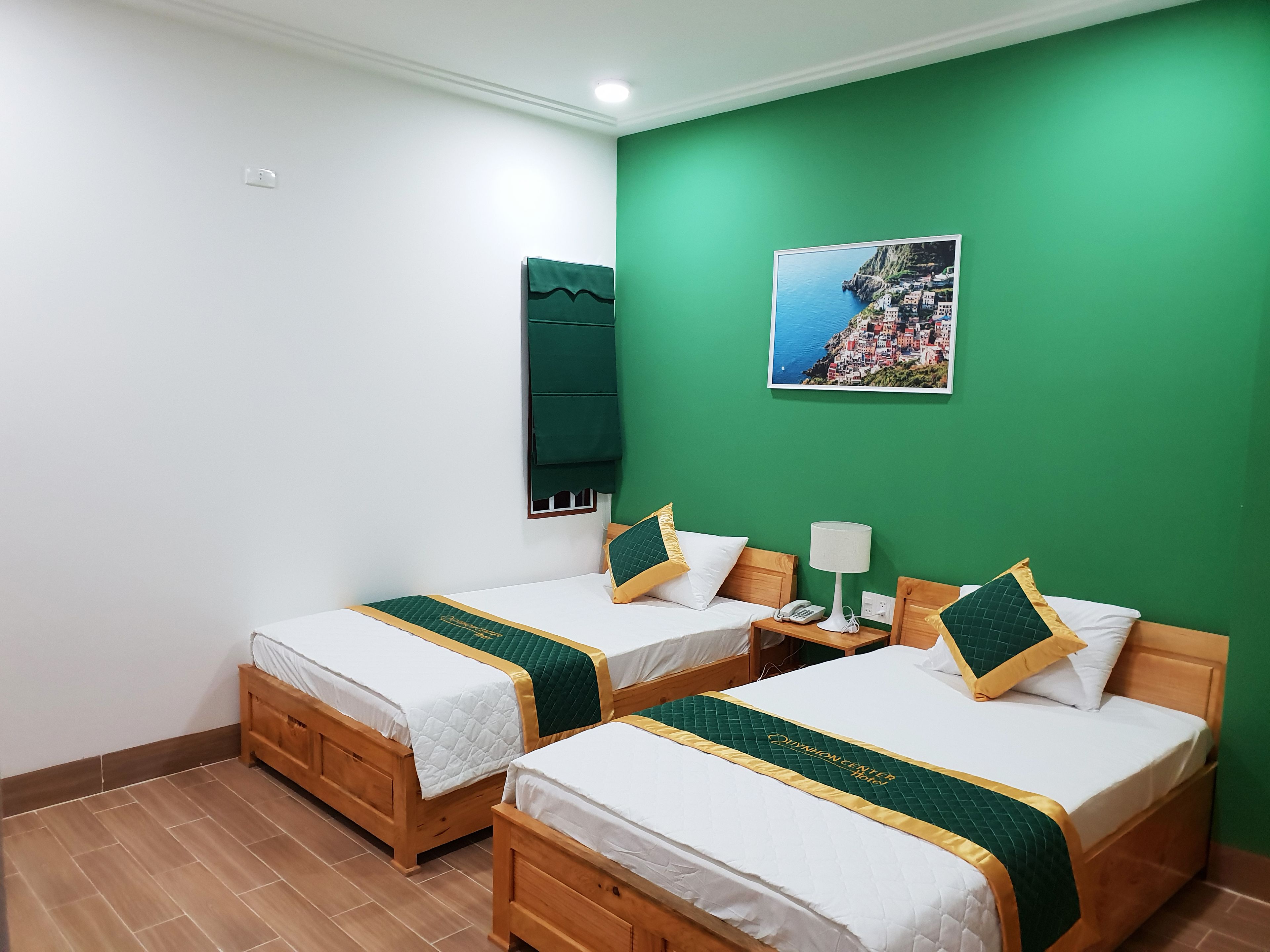 A hotel room with two wooden beds, green and gold bedspreads, a nightstand with a lamp, and a painting of a coastal town on the green wall.