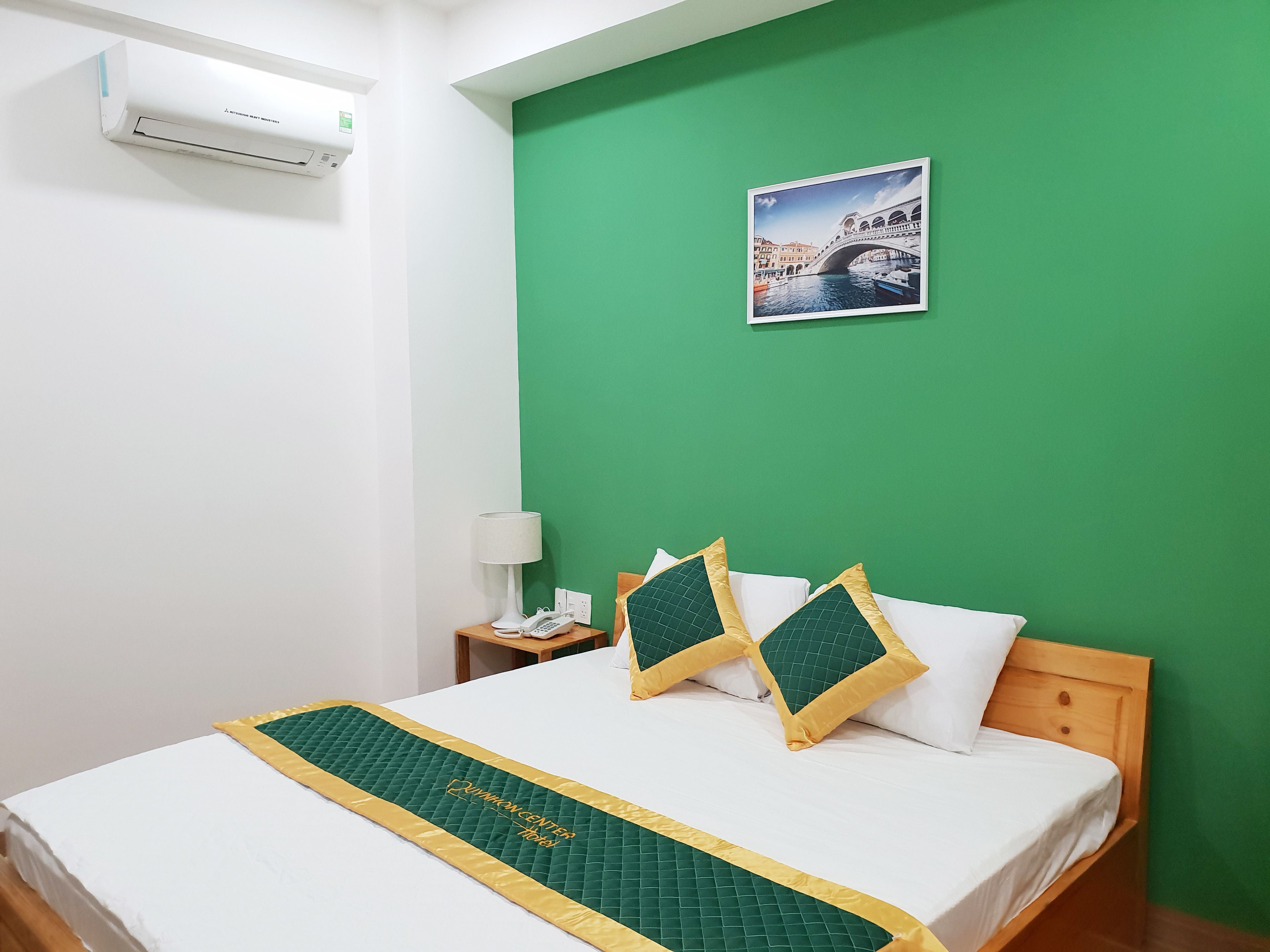 Habitación de hotel con pared verde, cama con cubrecama acolchado, dos almohadas, mesita de noche con lámpara y teléfono, y un cuadro enmarcado en la pared.