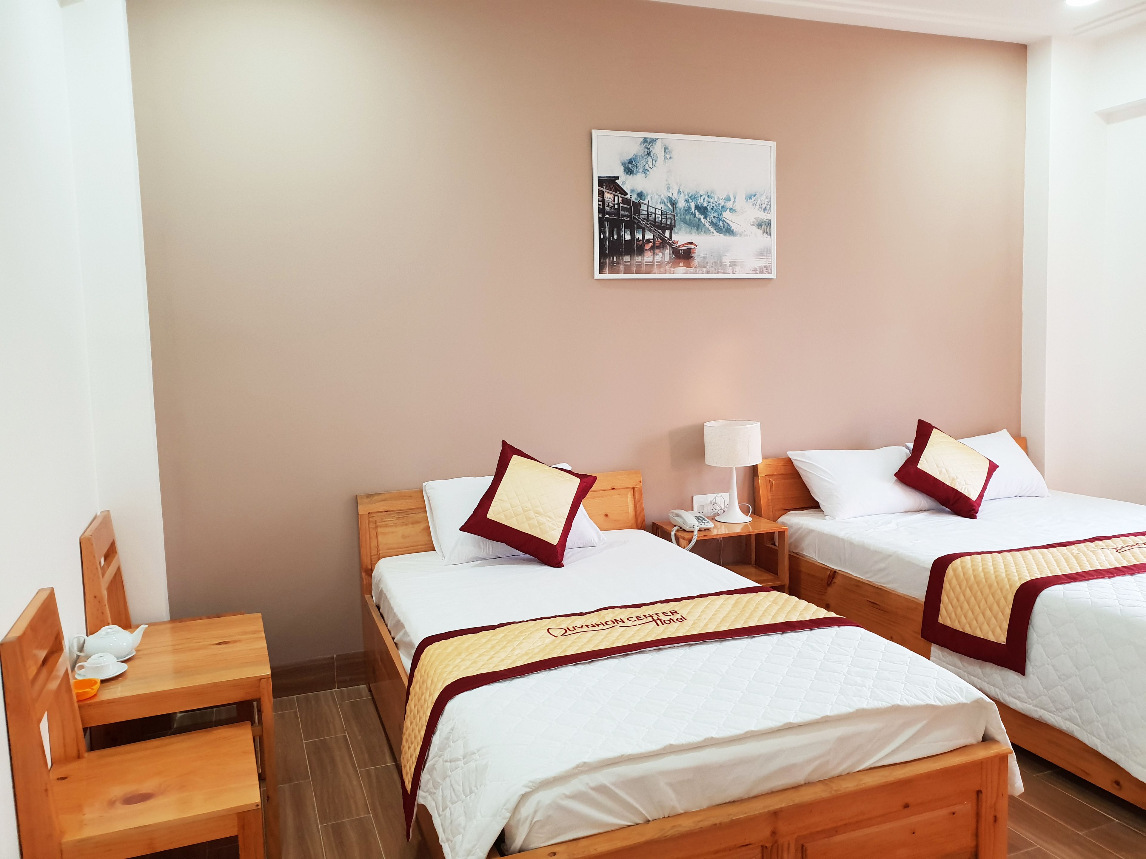 Een hotelkamer met twee bedden, houten hoofdeinden, een houten stoel, een tafel met theepot en een nachtkastje met lamp, en een schilderij aan de muur.