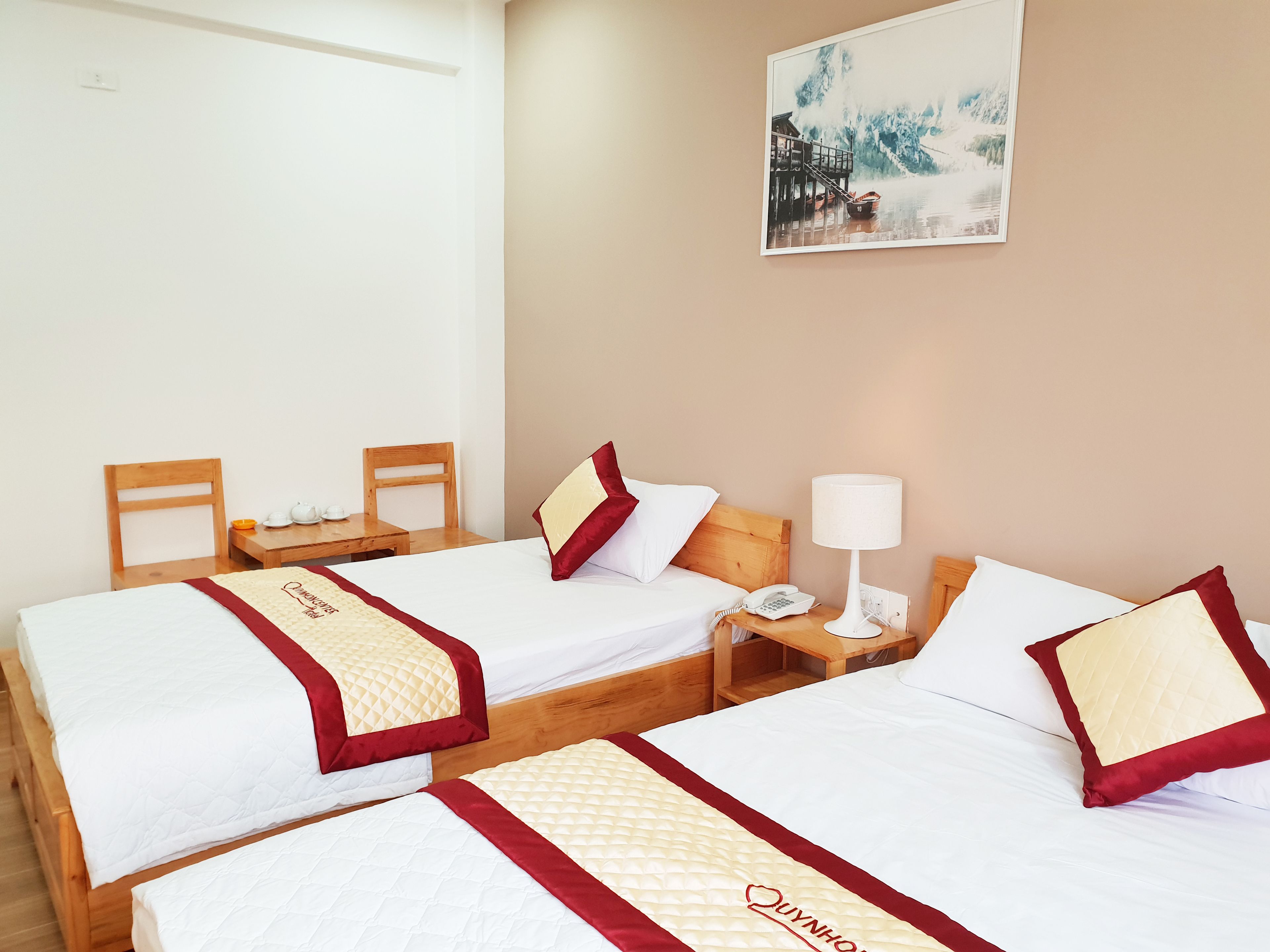 A hotel room with two single beds, each with a wooden headboard, white bedding, and red accents. There is a wooden nightstand with a lamp and a telephone. A painting of a coastal scene hangs on the wall.