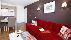 A modern living room with a red sofa, a dining area with a table and chairs, a kitchen with a stove, and a framed picture on the wall. - Cozy Chalet for You & Tails (Auris)