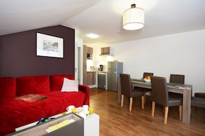 A compact living space with a red sofa, a dining table with chairs, a kitchenette, and a ceiling light fixture.