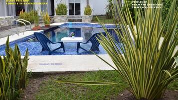 Una piscina con dos sillas, rodeada de plantas y una casa al fondo.
