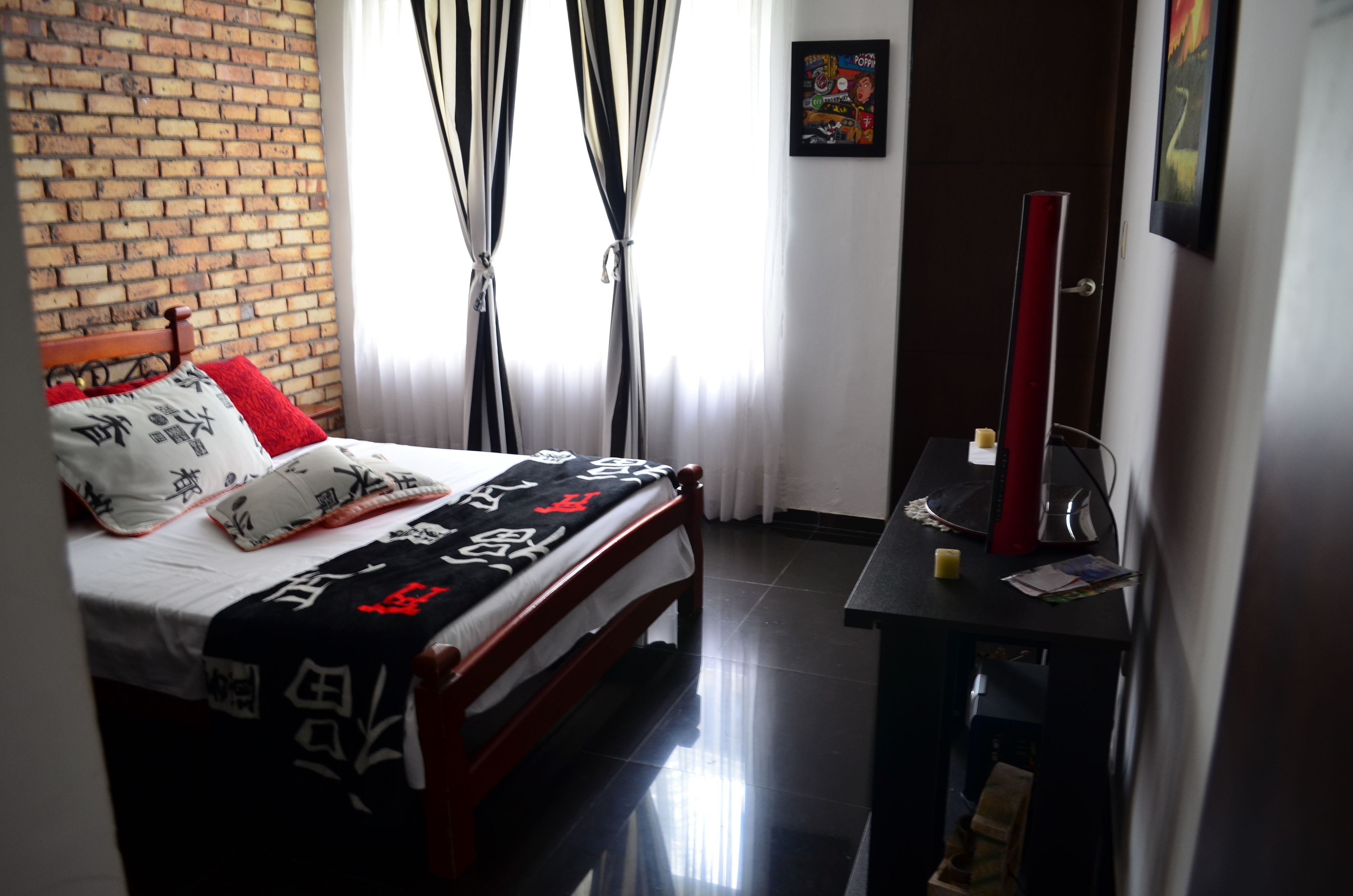 Una habitación de hotel con una pared de ladrillo, una cama con almohada roja y blanca, una colcha blanco y negro, un escritorio con espejo y un cuadro en la pared.