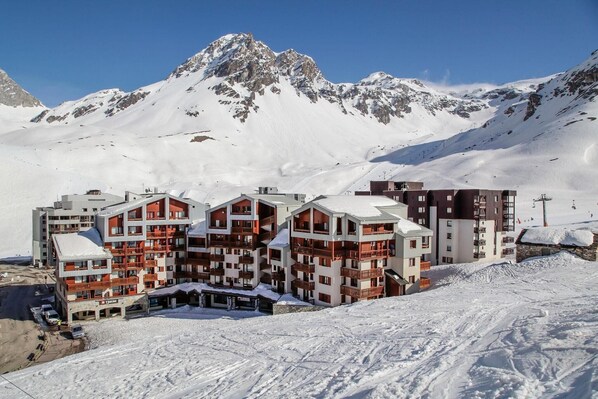 A snow-covered mountain resort with multi-story buildings featuring balconies and ski slopes in the background.