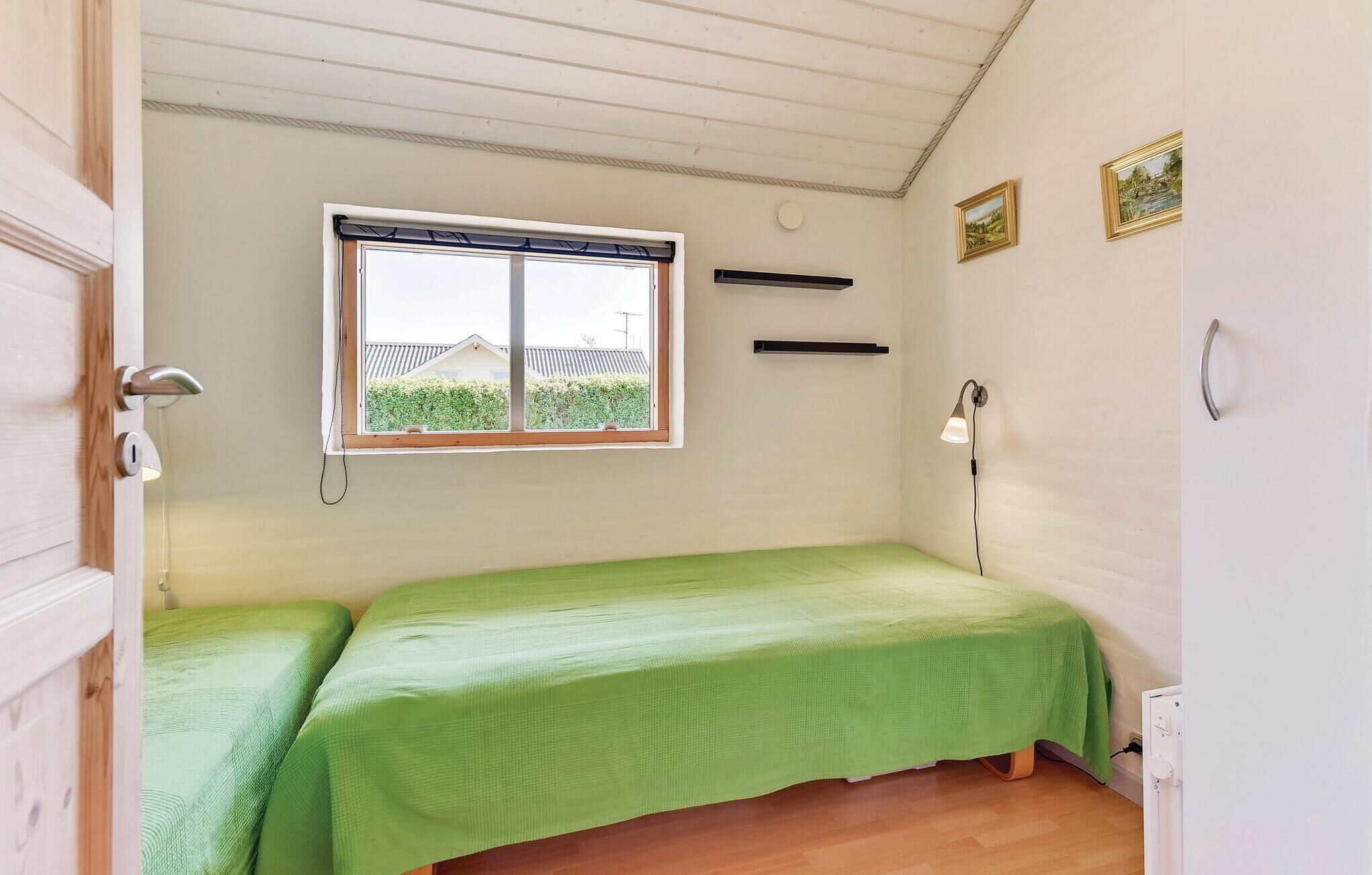 A small, single-bed room with a green bedspread, a window with a view of a house, a wall-mounted shelf, and a wall lamp.