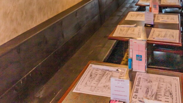 A restaurant interior with wooden booths, hanging pendant lights, framed artwork on the walls, and menus on the tables. - Historic Hotel Broadalbin (Broadalbin)