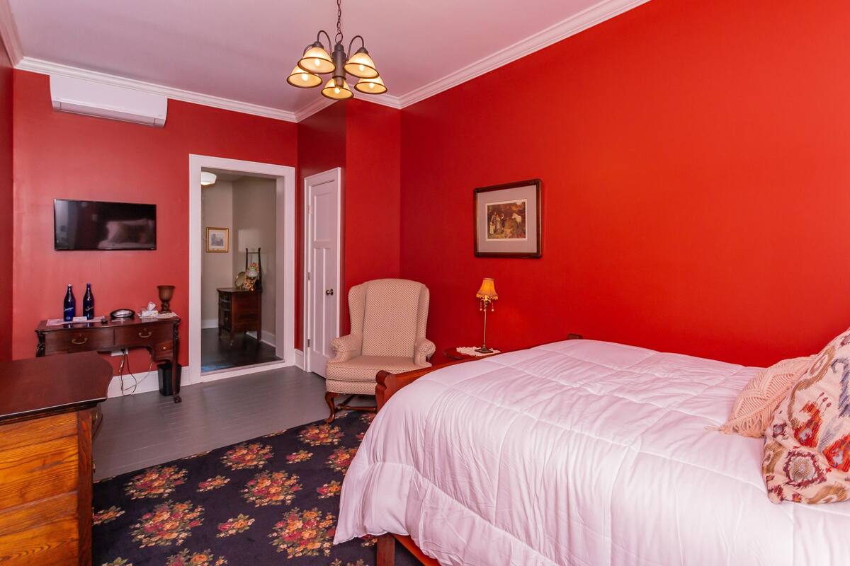 Ein Schlafzimmer mit roten Wänden, einem weißen Bett, einem floralen Teppich, einer Holzkommode, einem Stuhl und einem Schreibtisch mit Flaschen und einer Lampe.