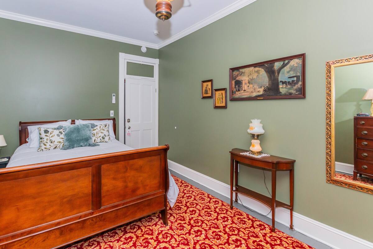 Ein Schlafzimmer mit einem hölzernen Bett, einem roten Teppich mit Muster, einer weißen Tür, einem kleinen Tisch mit Lampe und einem großen Spiegel mit aufwendigem Rahmen.
