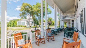 A covered porch with wooden rocking chairs and tables, overlooking a street with a white building in the background. - Historic Hotel Broadalbin (Broadalbin)