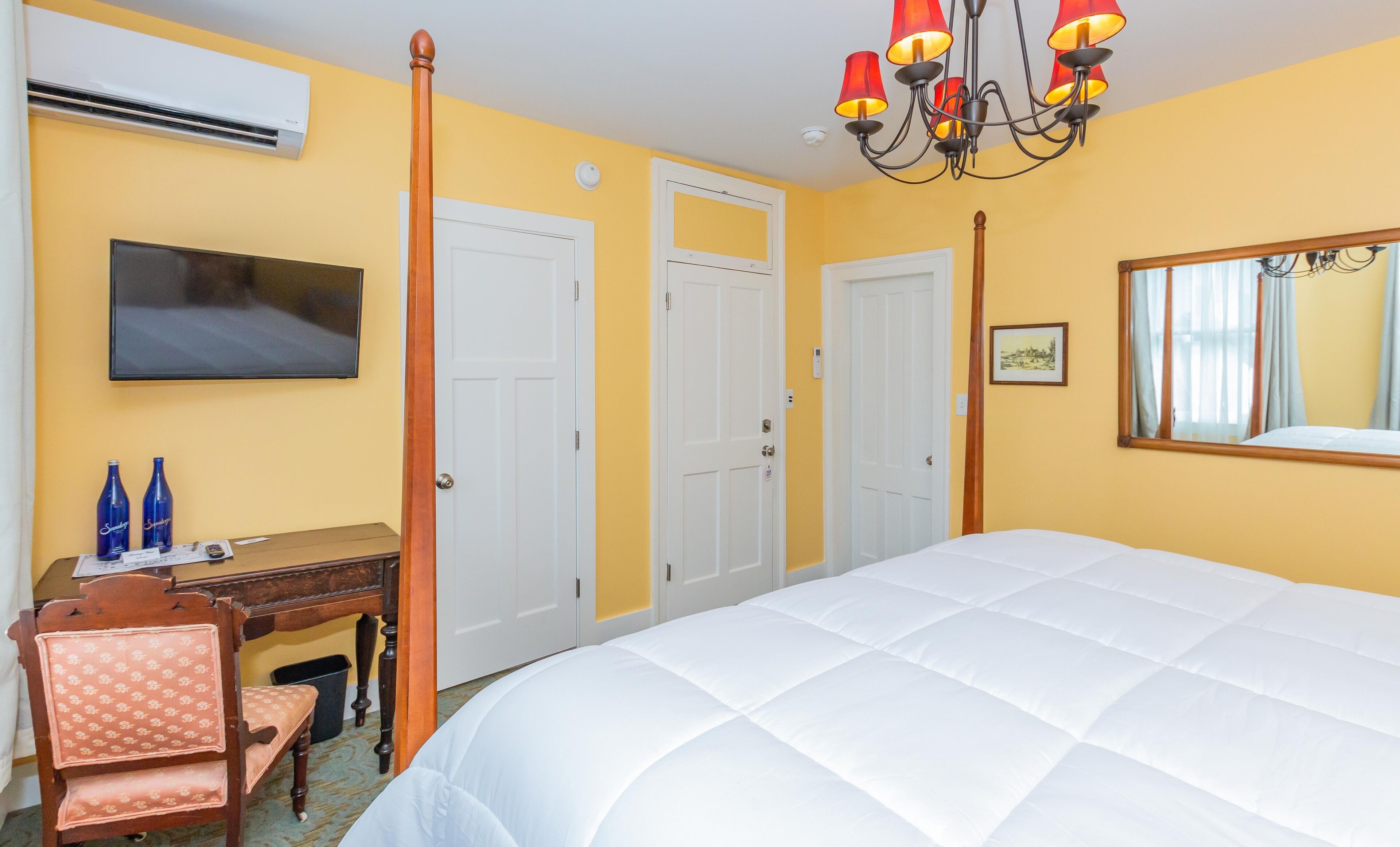 A hotel room with a large bed, a desk with two blue bottles, a chair, a television, and a chandelier.