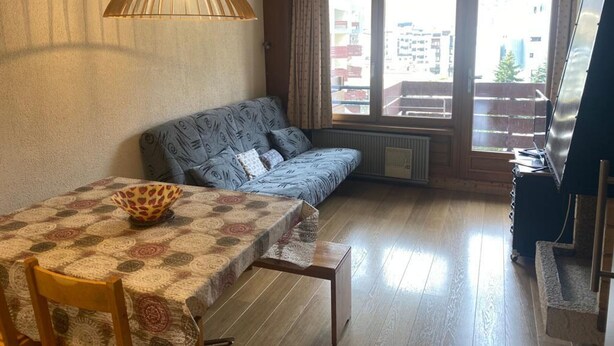 Ein gemütliches Wohnzimmer mit einer Couch, einem Esstisch mit gemustertem Tischläufer und einem großen Fenster mit Blick auf Gebäude.