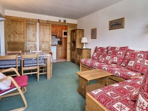 A cozy room with a wooden sofa, a dining table with chairs, a small wooden table, and a kitchen area with wooden cabinets and a stove.