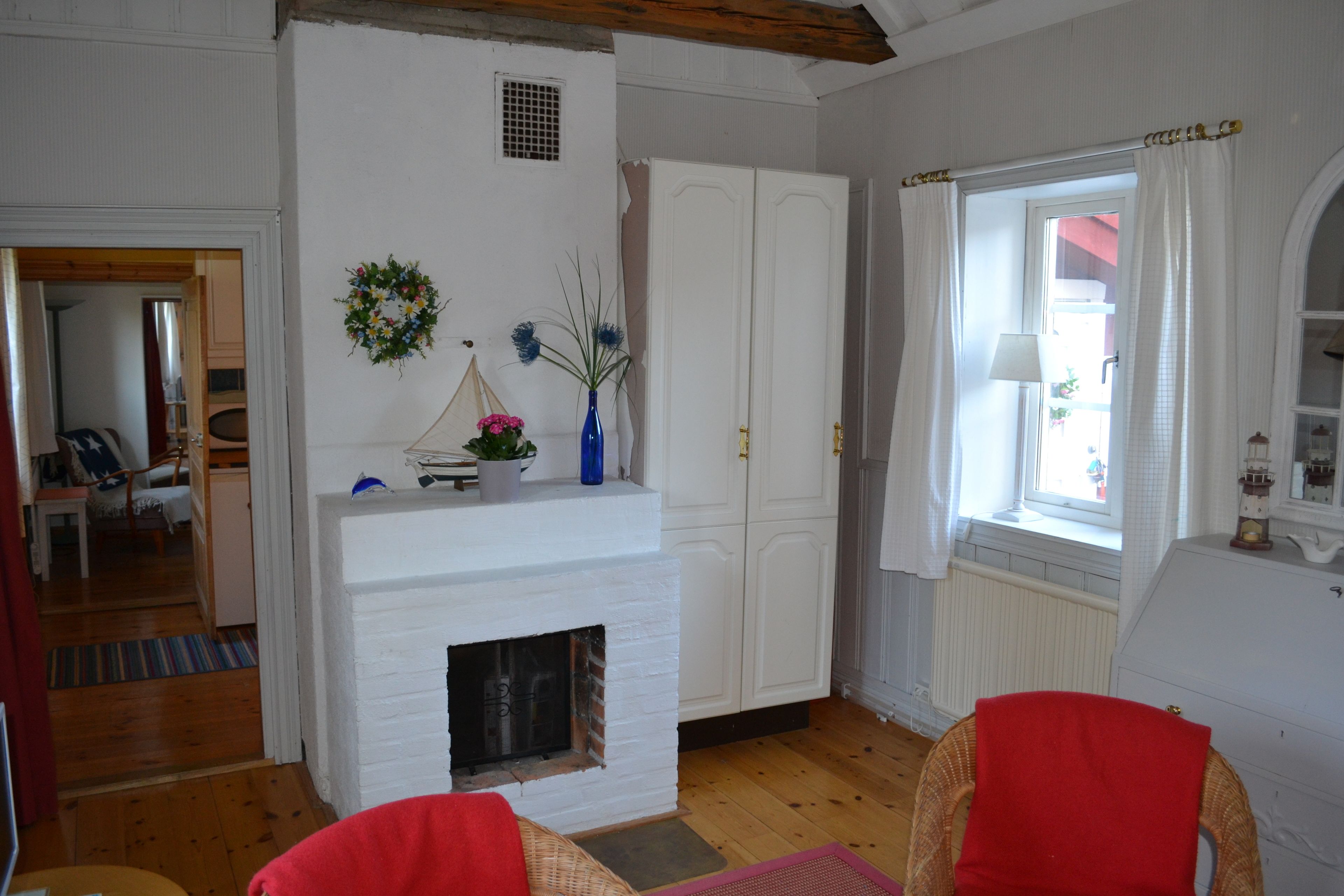 A room with a white fireplace, wooden flooring, and white walls. There is a window with white curtains and a view of the outside.