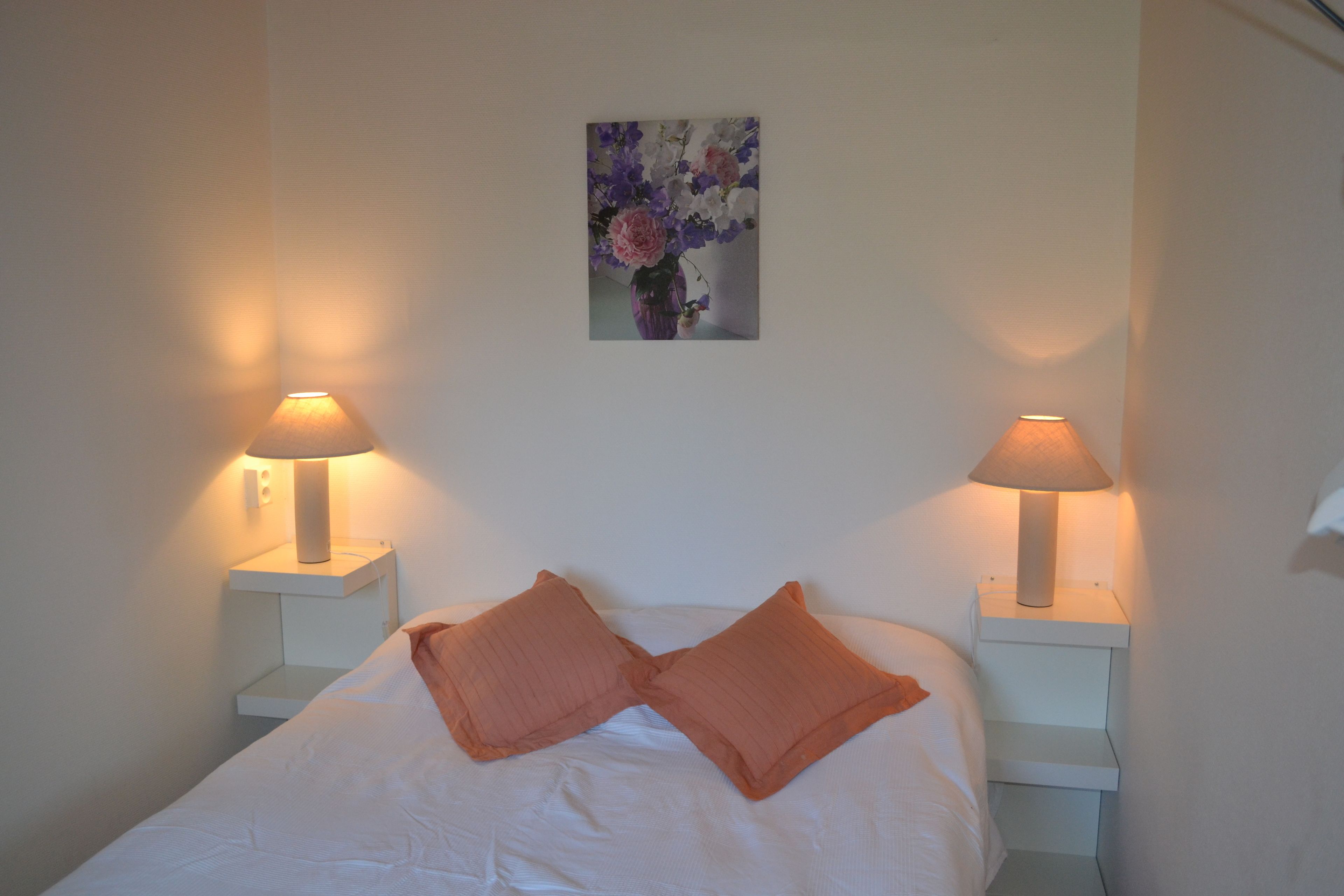 A neatly made bed with two pillows, two bedside lamps, and a framed floral artwork on the wall.