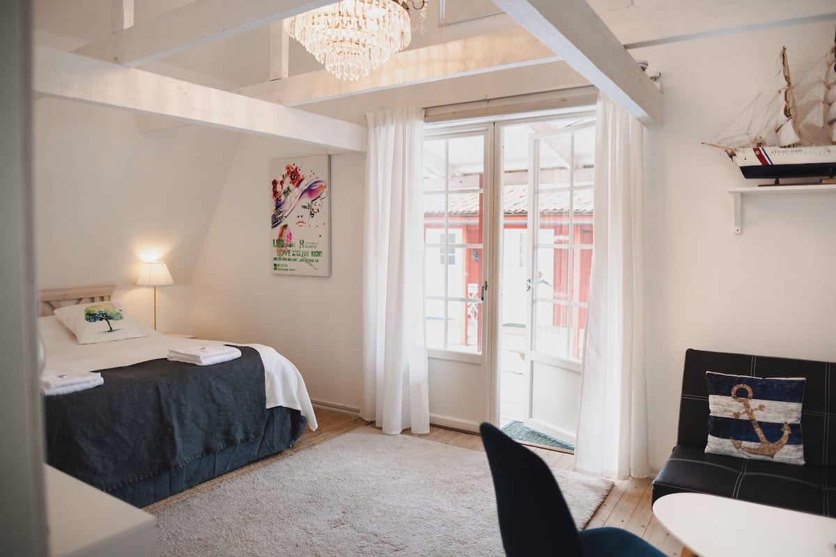 A bedroom with a bed, a desk, a chair, a chandelier, and a view of the outside.