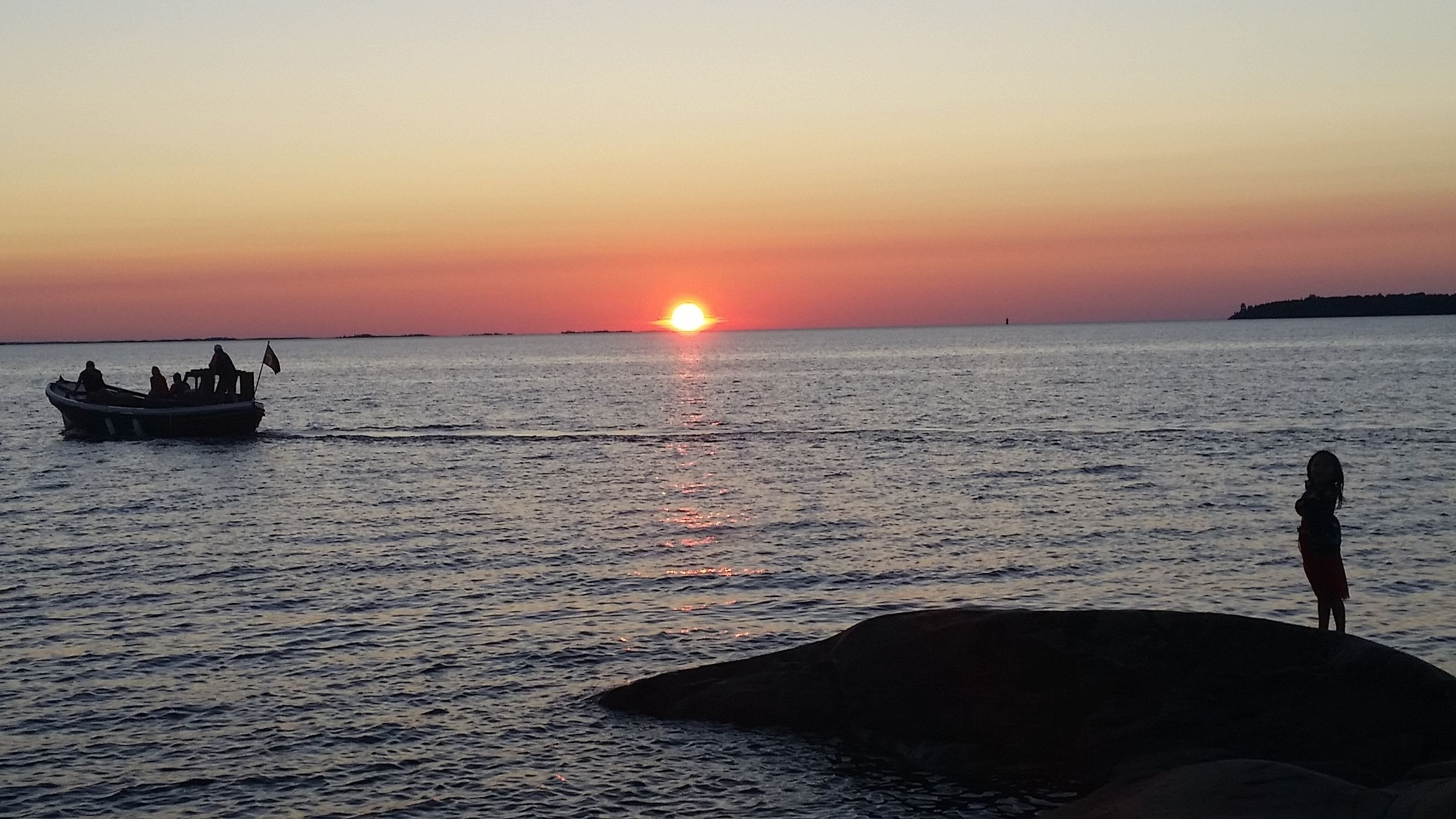 Een zonsondergang boven een rustige zee, met een boot en een persoon die op een rots staat.
