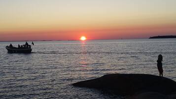 Een zonsondergang boven een rustige zee, met een boot en een persoon die op een rots staat.