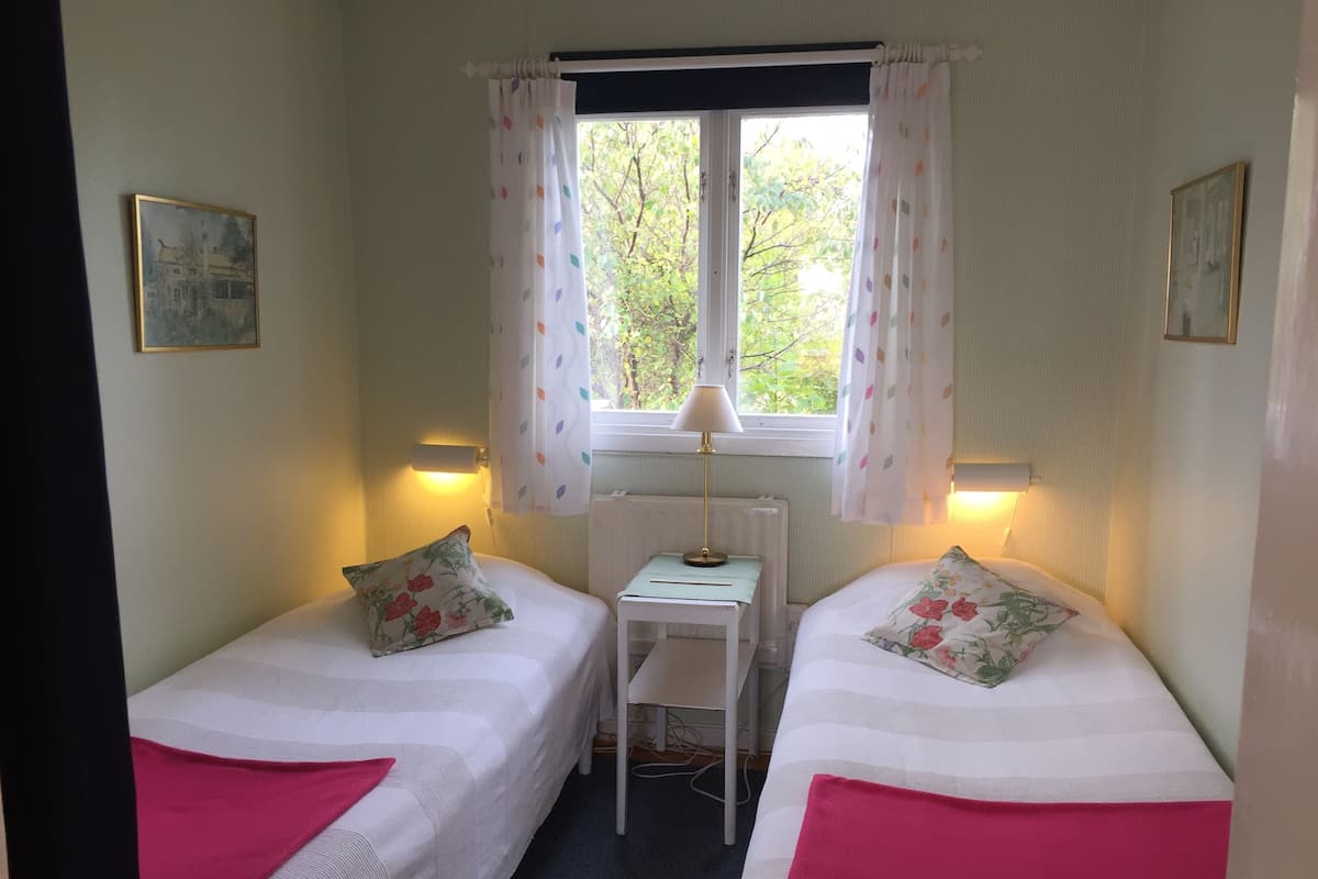 A room with two beds, a small table, a chair, a window with curtains, and wall-mounted lamps.