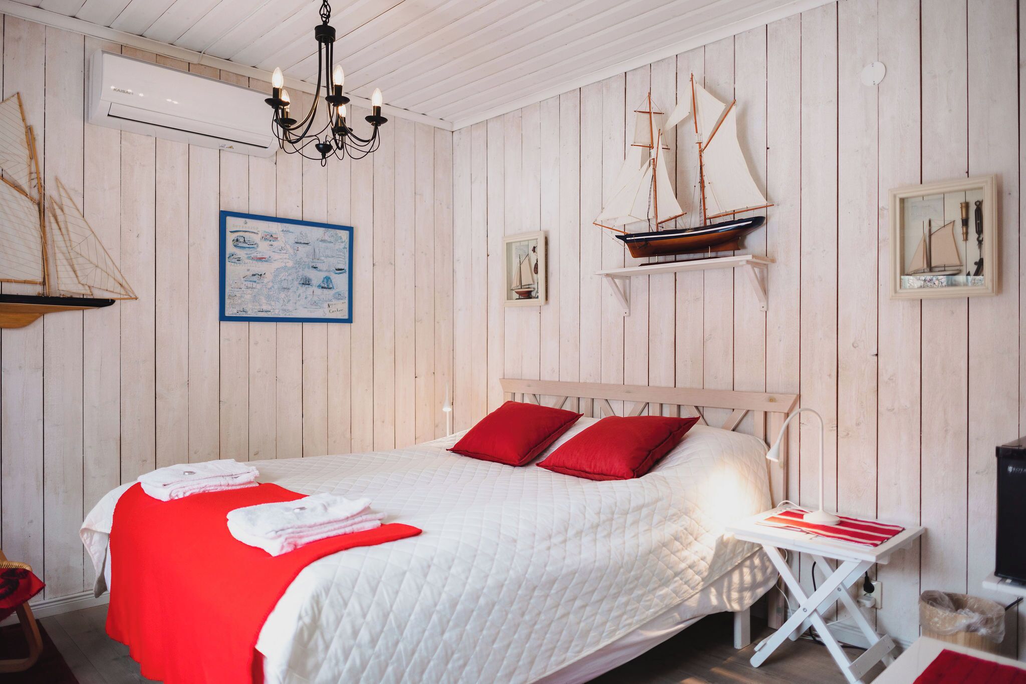 Un dormitorio con un panel de madera en la pared, una cama con ropa de cama blanca y roja, una mesita de noche con una lámpara y un modelo de barco.