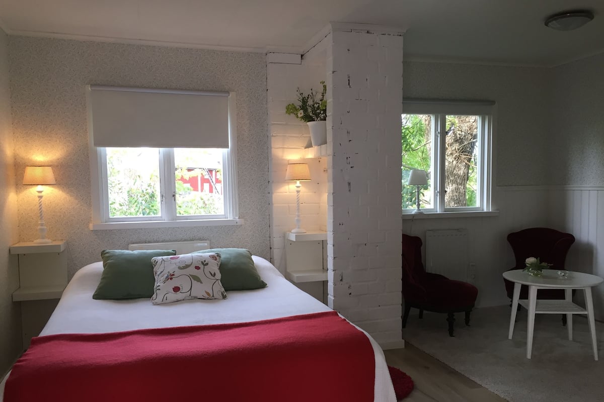 A bedroom with a bed, two windows, a chair, a table, and a radiator.