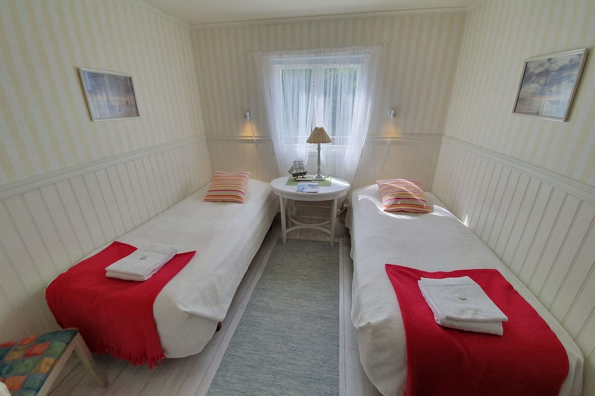 A small, cozy room with two single beds, a small table with a lamp, and a window with sheer curtains.