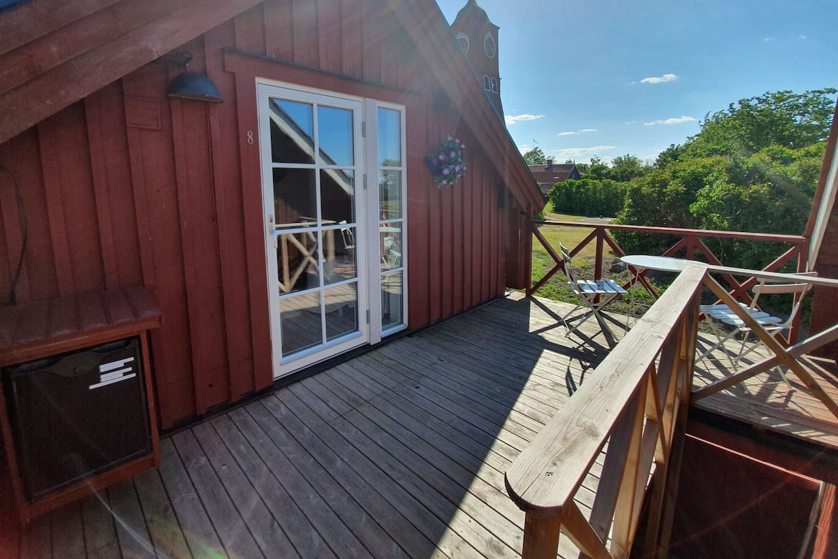 A wooden deck with a glass door leading to an interior space, surrounded by a railing and a view of greenery.