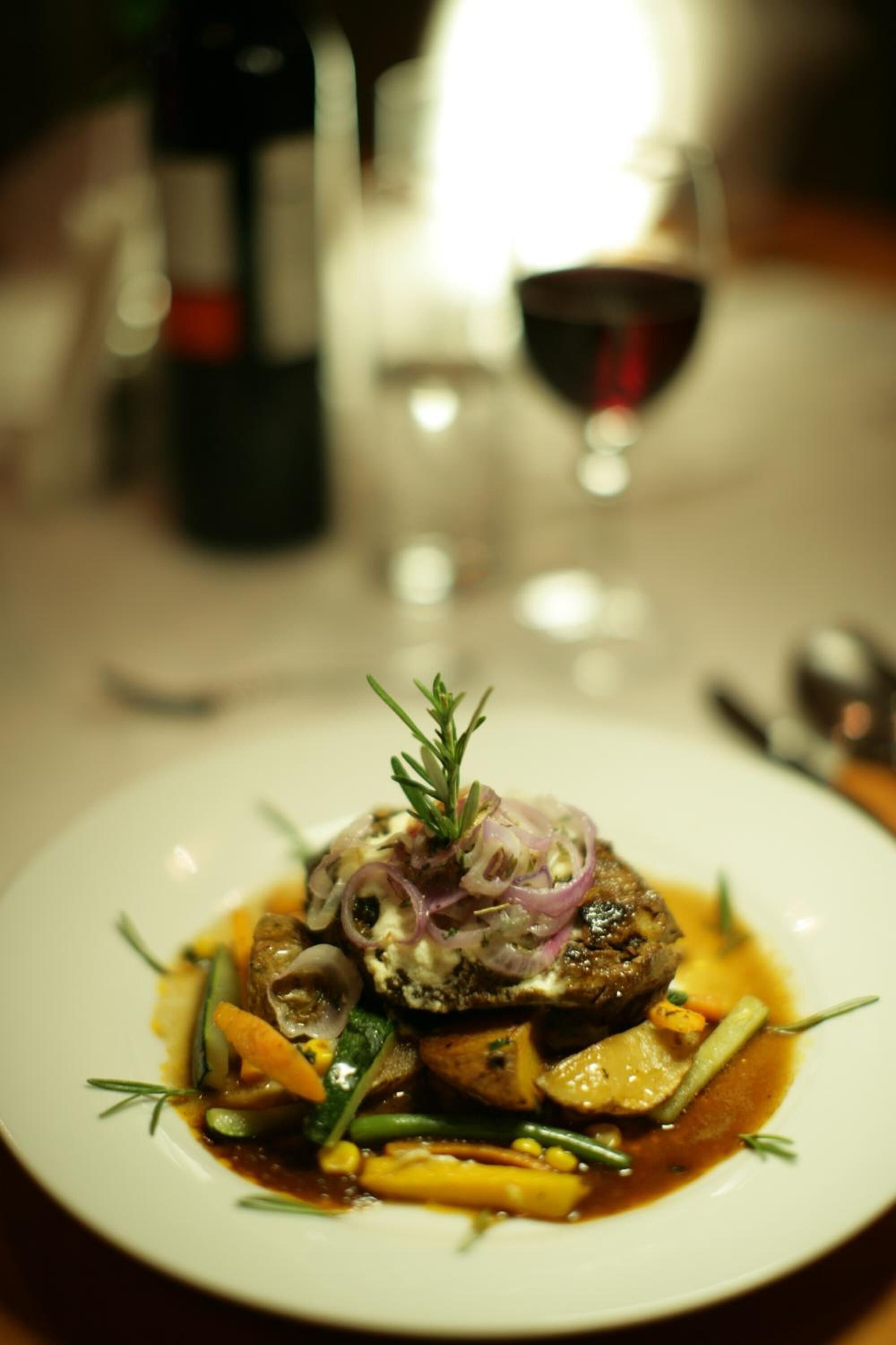 Un plato de comida con guarnición de hierbas y cebollas, acompañado de una botella de vino y una copa de vino tinto al fondo.
