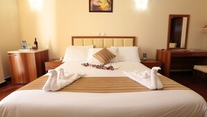 A hotel room with a large bed, a wooden nightstand with a lamp, a desk with a chair, and a painting on the wall. - Bete Daniel Hotel (Bahir Dar)