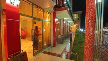 L’entrée d’un hôtel, avec une porte en verre, un pilier rouge et un petit espace extérieur aménagé avec des sièges, offrant une vue sur un jardin.