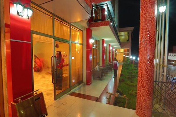 Entrada de un hotel con puerta de vidrio, un pilar rojo y una pequeña zona de estar al aire libre con vista a un jardín.