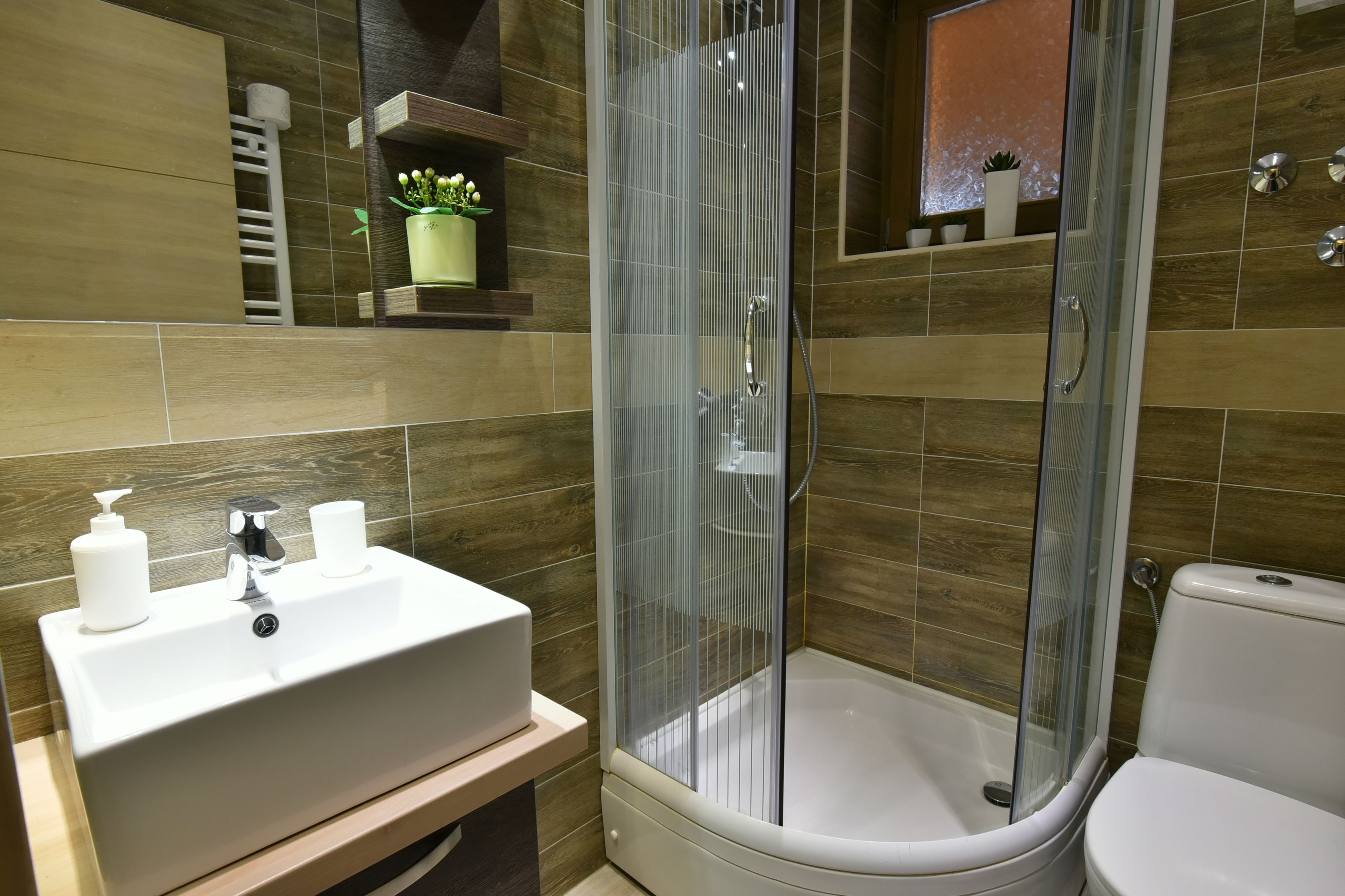 Un baño moderno con ducha envuelta en vidrio, lavamanos blanco con dispensadores y un inodoro.