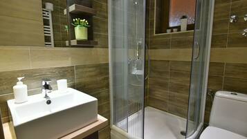 Un baño moderno con ducha envuelta en vidrio, lavamanos blanco con dispensadores y un inodoro.