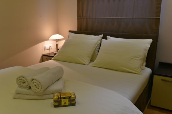 Una cama bien hecha con sábanas y almohadas blancas, una mesita de noche con una lámpara, y una caja de artículos de aseo.