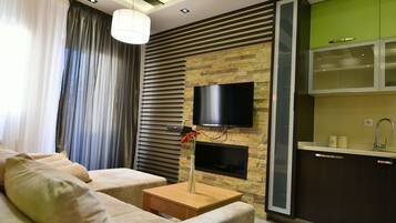 A modern living room with a sofa, a side table, a vase with flowers, a flat-screen TV mounted on a stone wall, and a kitchen area with green cabinets and a sink.