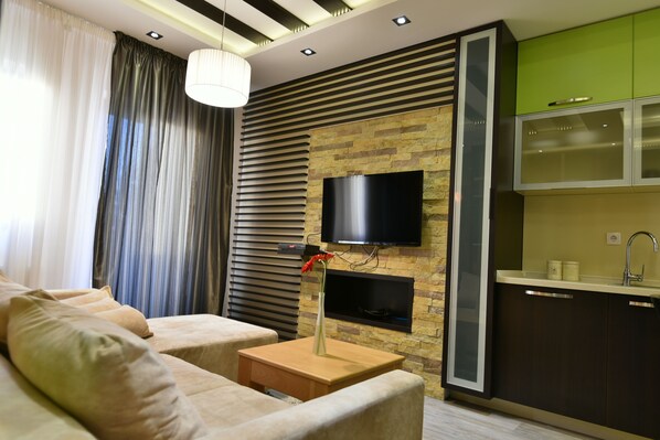 A modern living room with a sofa, a side table, a vase with flowers, a flat-screen TV mounted on a stone wall, and a kitchen area with green cabinets and a sink. - Villa Natural Wood (Zlatibor)