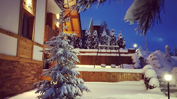 Un espacio exterior cubierto de nieve con un edificio de madera, un árbol nevado y una escalera que sube a un nivel superior.