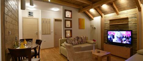A cozy living room with a wooden ceiling, a dining area with a table and chairs, a sofa, and a television.