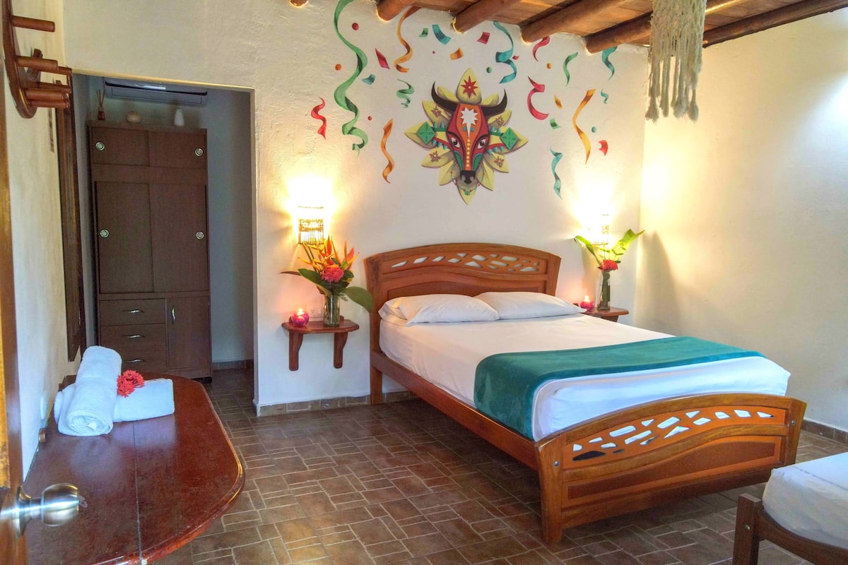 A bedroom with a wooden bed, a nightstand with a lamp, a wall-mounted shelf with a vase of flowers, and a door with a towel on the floor.