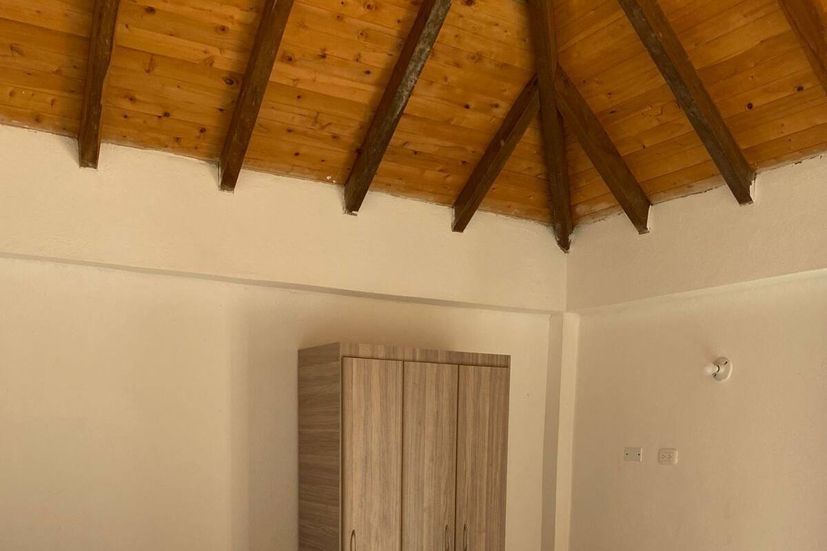 A bedroom with a wooden ceiling, a bed with white bedding, a green throw blanket, and a wooden wardrobe.