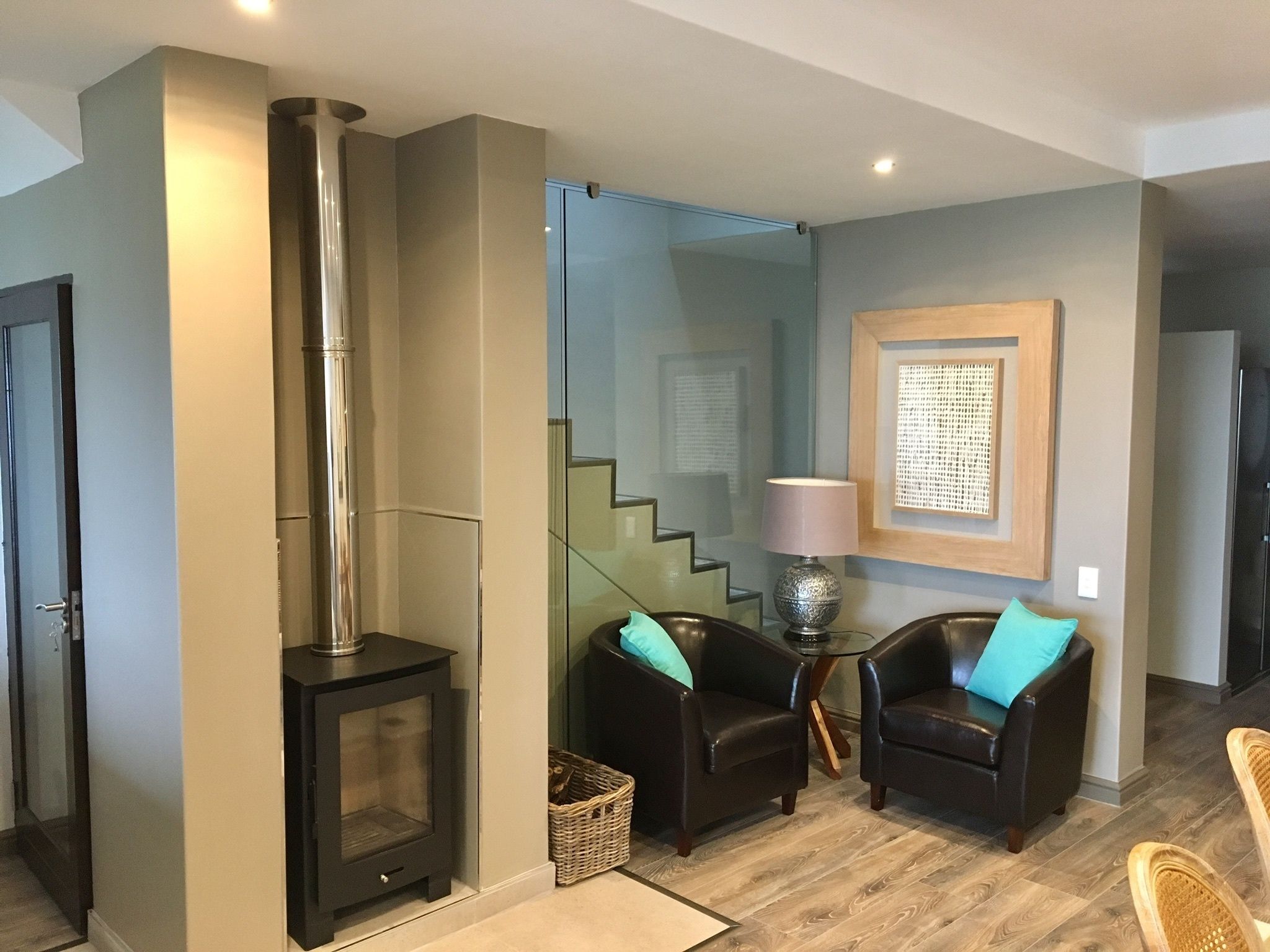Un salon moderne doté d’un poêle à bois, de deux fauteuils en cuir avec des coussins turquoise, d’un escalier en verre et d’une œuvre d’art encadrée accrochée au mur.