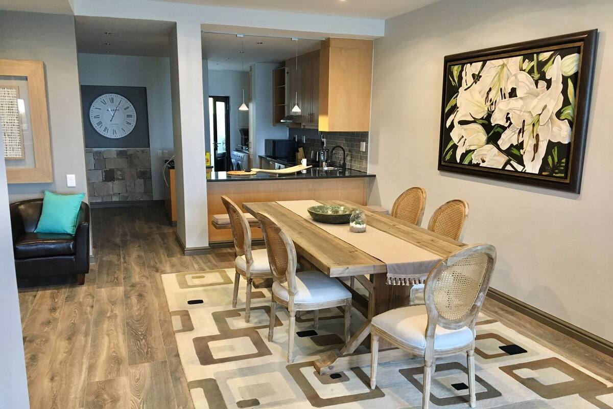 A modern kitchen with wooden cabinets, a dining area with a wooden table and chairs, and a large clock on the wall.