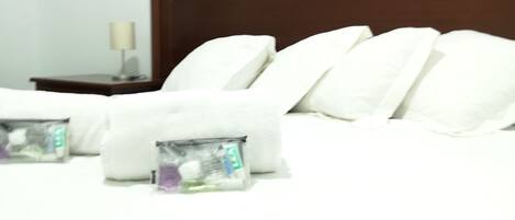 Un lit bien fait, avec du linge de lit blanc et des oreillers, une tête de lit en bois et une table de chevet sur laquelle se trouvent une lampe et une petite boîte contenant des articles de toilette.