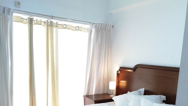 Habitación de hotel con una cama grande, ropa de cama blanca, cabecero de madera, una mesita de noche y una ventana con cortinas.